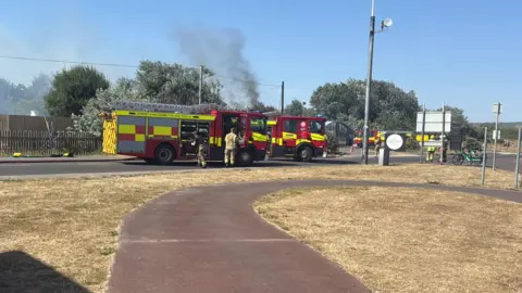 Fire engines parked up along a road with smoke rising in the background