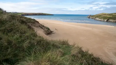 A wide sandy beach with a few people walking along the shore, bordered by grassy dunes and cliffs. The ocean stretches into the background with gentle waves, and the sky is partly cloudy with some blue patches visible.