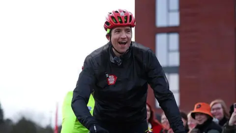 Greg James smiles as he rides past a group of supporters cheering him.