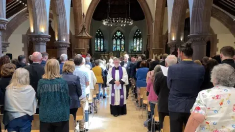People standing in the cathedral with a member of the clergy walking down the middle aisle. The building has arches and stained glass windows.
