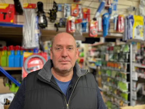 Paul Crone wearing a black gilet standing in his hardware store with bottles of spray and tools behind him on shelves 