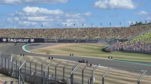 Kris Holland/BBC A wide view of the Landostand at Silverstone Circuit. Max Verstappen is shown driving his Red Bull in the foreground.