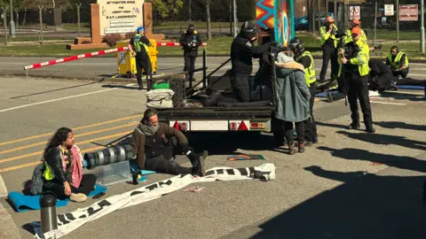 Demonstrators sitting on the floor and attached to a vehicle outside the entrance to RAF Lakenheath. Police officers are also on the scene. 