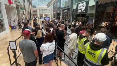 Men and women are queued up near railings outside the Oasis Live '25 pop-up shop. Two men are wearing yellow high-visibility vests and one has a hard hat. A sign directs people where to queue. The Bullring shopping centre can be seen in the background.
