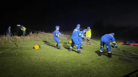 People wearing blue overalls using a rope pully system on grass at the top of a cliff