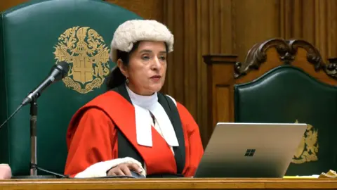 PA A female judge dressed in red robes and a wig. She has dark hair, tied back and is sat at a wooden desk with a Microsoft laptop in front of her.