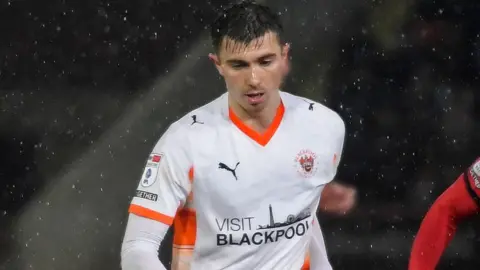 Scott Banks wearing a white Blackpool kit playing on a rainy evening