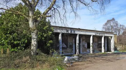 White single-storey building with "RAF Upwood" in black letters above the entrance. The facade has six pillars running from floor to roof. There is graffiti on the pillars. Wooden boards are visible behind the pillars. There are trees either side of the building. 