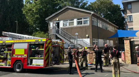 A fire engine and a large group of firefighters stood outside the large brown nursery building.