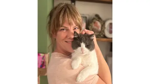 Kat Steele A smiling woman holds her mischievous cat who has been stealing cat food pouches from one of her neighbours.