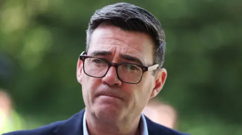 A middle aged man with dark, slightly greying hair. He is wearing a dark blue suit jacket, a white shirt and dark , squarish rimmed glasses. His lips are pursed and he has a serious look.