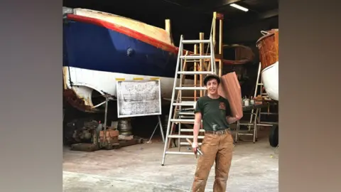 Contributed A woman stands inside a building in a boat yard. She is smiling at the camera as she holds a large piece of wood in one hand and rests it on her shoulder. She is wearing a flatcap, green T-shirt and brown trousers. She is holding some tools in her other hand, There are boats raised on contraptions behind her.