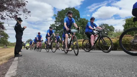 People on bicycles cycling on a road and wearing blue cycle jerseys. A female police officer is at the side of the road in her uniform applauding the cyclists.