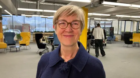 Judith Squires looks at the camera. She has short blonde hair and glasses. She wears a navy blue jacket. 