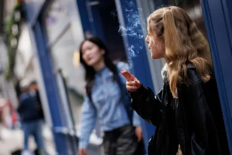 A woman with long, wavy blonde is standing outside smoking a cigarette. Behind her, another woman walks out through a doorway.