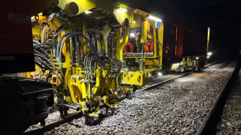 Submitted A large, bright yellow machine is on train tracks in the dark. It is lit by lights on the machine and people operating it are wearing hi-vis orange clothing and white hard hats.