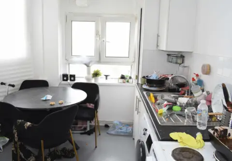 PA Media A kitchen with a round table, a cluttered surface above appliances, and a double window