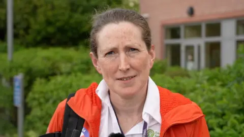 Amy Heys looks at the camera. She has brown hair pulled back in a pony tail, is wearing a white shirt with a green Environment Agency logo on the breast and an orange mountain rescue jacket. Behind her is a green hedge