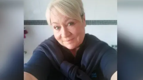 Family photo Kelly smiles as she takes a selfie. She has short blonde hair swept to one side and is wearing a dark blue top. Behind her is a plain cream wall