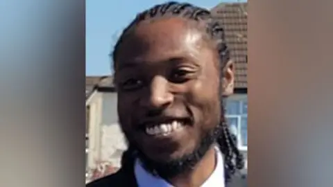 Met Police Jordan Briscoe, who is wearing a white shirt and black jacket, stands in front of a house