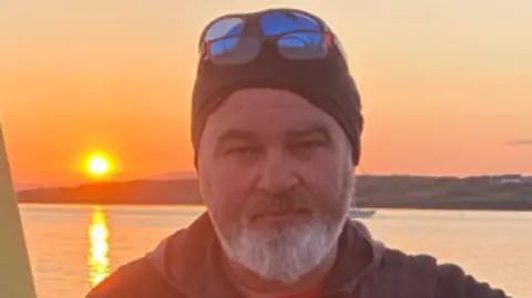 Handout Shows a man with a woollen hat and glasses above his head with sea in the background and sun reflecting on the water