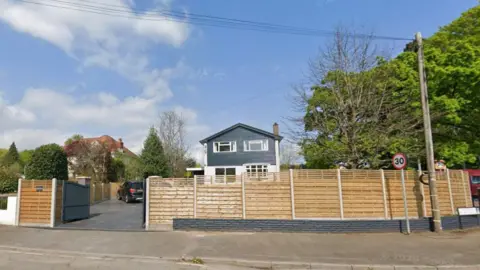 A house on St Lawrence Road, Chepstow. It has a tall timber fence around the edge and a grey slate-style driveway with a dark-coloured SUV parked on it. The house is two storeys and is white on the bottom half with a dark, panel style for the top half. Large trees stand to the right of the house 