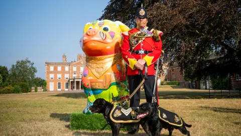 Staffordshire County Council A man wearing a red ceremonial military uniform is standing alongside a brightly-coloured sculpture of a Staffordshire bull terrier. The man has two dogs with him on leads. In the distance is a historic country house.