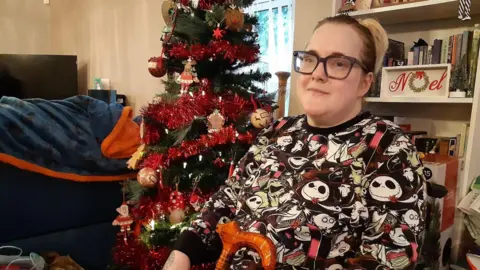 Charlotte Hale is sitting in her living room with a bookshelf behind her, which has a decorative sign saying Noel, and there is a Christmas tree with red tinsel and baubles. Miss Hale is wearing glasses and holding a walking stick.