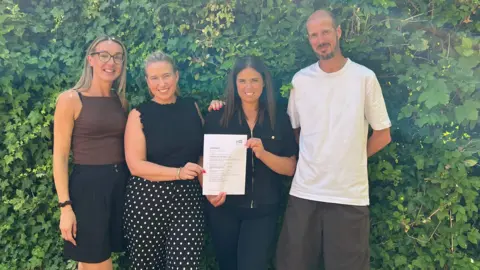 The image shows four people smiling and posing for the camera, while the two people in the middle hold up a certificate.
