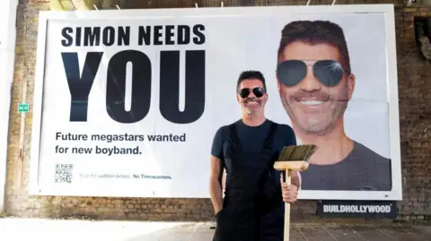PA Media/David Parry Simon Cowell is wearing sunglasses, black dungarees and a blue T-shirt and is smiling while holding a wooden broom. Behind him is a billboard with a photo of him smiling and 'Simon needs you. Future megastars wanted for new boyband'.