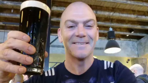 Mark Hehir, a bald man smiles, as he holds a Guinness in his hand