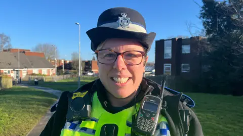 Katy is pictured looking into the camera and smiling. She's wearing police uniform; a black jacket over the top of a yellow hi-vis vest, with another black top underneath that. She's wearing a black police bowler hat with the West Mercia Police badge on it. On her right shoulder she's got a police radio, and on her left shoulder a body-worn camera. She's stood on a footpath on the Brookside estate in Telford, and in the distance you can see red brick flats, houses, and bungalows. Closer to the camera there's grassy spaces and trees. The sky is bright blue and clear, with lots of sunshine. 