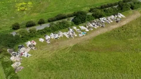 Aerial view of a long line of fly‑tipped rubbish dumped on a strip of land beside a rural road.
