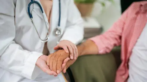 A doctor holds a woman's hand in a consultation. The doctor has a white coat and a stethoscope.