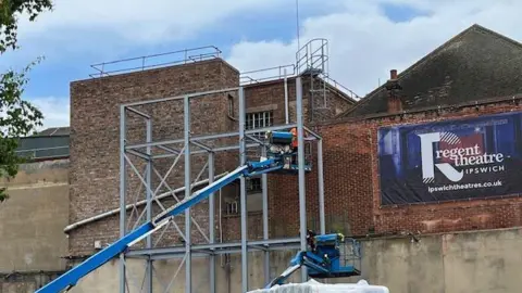 Ipswich Borough Council Scaffolding is placed against the external part of the theatre. A builder can be seen on top of the scaffolding frame working on the building. The theatre's sign can be seen on part of the wall to the right of this.