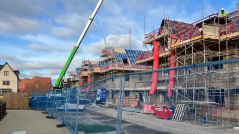 A metal fence with three-storey houses being built behind it, with scaffolding around it. There is a crane, blue skies and white clouds above.