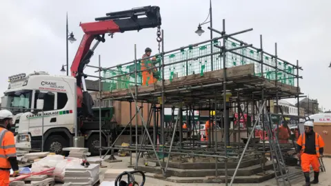 John Devine/BBC An orange and black crane arm is lifting sections of the fountain into place with scaffolding shrouding the site, there are three stone steps leading to the base of the structure and there are several workmen in high-viz orange outfits and hard hats on top of the 3m (nearly 9ft) high scaffolding and some below.