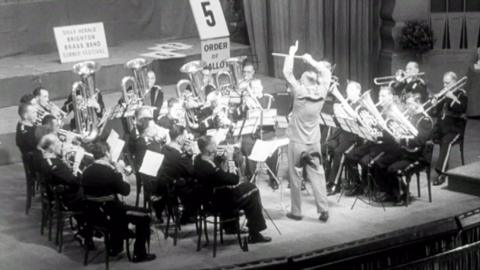 Brass band, led by conductor, play music on stage.