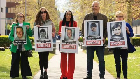 PA Media Five people stand in a line holding placards of the shooting of five people in west Belfast in 1972