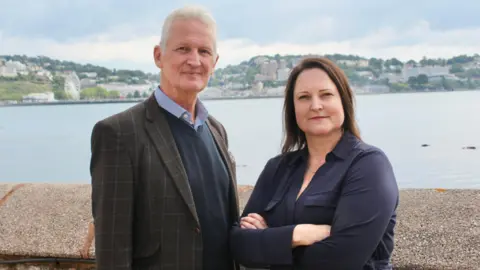 Mark Kingscote and Alison Hernandez are smiling at the camera as they stand in front of a harbour wall with the sea behind them and houses visible on land in the distance. He is wearing a checked jacket over a blue jumper and shirt and she is wearing a navy blouse and has her arms crossed.