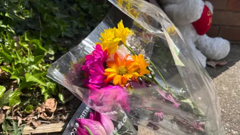 Flowers, purple, yellow and orange, are in the middle of the photo. A mainly white cuddly toy is on the right.