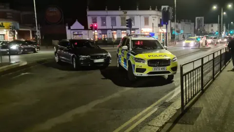 A police car and a black Volvo estate are parked in the middle of a road that appears to have been closed off to traffic. It is night-time and there are people walking on the pavement at the side of the road that has been closed off. 