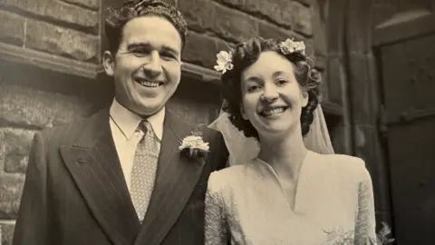 Ruth Pole A black-and-white photo shows a smiling couple on their wedding day. The man is on the left and has dark hair. He is wearing a pin striped suit with a carnation and a patterned tie. The woman is on the right and has curled dark hair, with flowers and a veil. She is wearing a white v-necked wedding dress.
