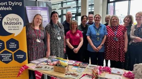 NHS Nine women and two men, many wearing name badges, stand behind a table covered in leaflets and pens. A pull-up stand reads "Dying Matters, it's time to talk about it".