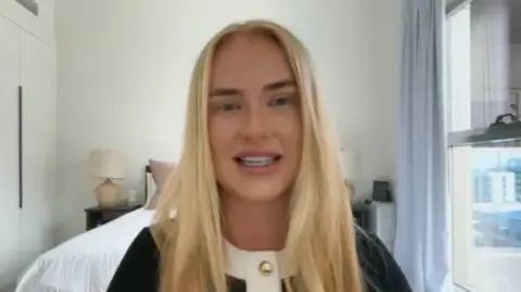 A woman with long blonde hair sits indoors in a bright, modern bedroom, speaking toward the camera. She is wearing a black top with a white collar and gold buttons. Behind her is a neatly made bed, bedside table with a lamp, built-in wardrobes, and a large window with curtains showing nearby buildings outside.