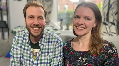 BBC A man with brown hair and a beard, wearing a checked shirt, smiles at the camera. Beside him is a woman with long brown hair and wearing a flowery dress, also smiling at the camera as they both sit on a grey sofa.