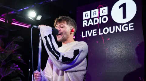 A man is singing into a microphone on a stage with a “BBC Radio 1 Live Lounge” sign in the background.