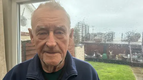 Robby West/BBC Cliff Williams has short grey hair and is wearing a green T-shirt and a navy jacket. He is standing in front of a window, with his garden behind him.