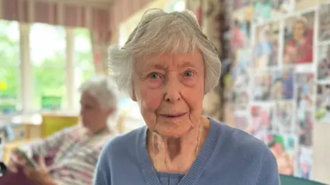 Alice Cunningham/BBC An elderly woman with short white hair smiles at the camera. She wears a blue jumper and gold necklace. Behind her there is another woman sitting in the room.