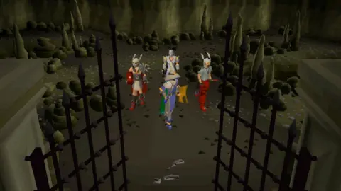 Jagex Four characters stand in front of an open gate in a cave, seemingly to enter part of a dungeon. The graphics are reminiscent of a video game from 25 years ago - very pixel-y. The characters are wearing different kinds of armour and one appears to be dressed like a wizard.